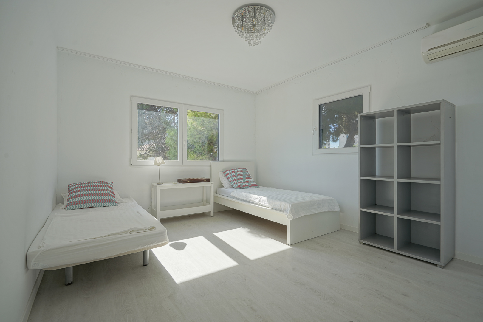 Dormitorio luminoso con dos camas individuales, suelos de madera clara y estantería gris. Incluye aire acondicionado, lámpara de cristal y ventanas con vistas al jardín.
