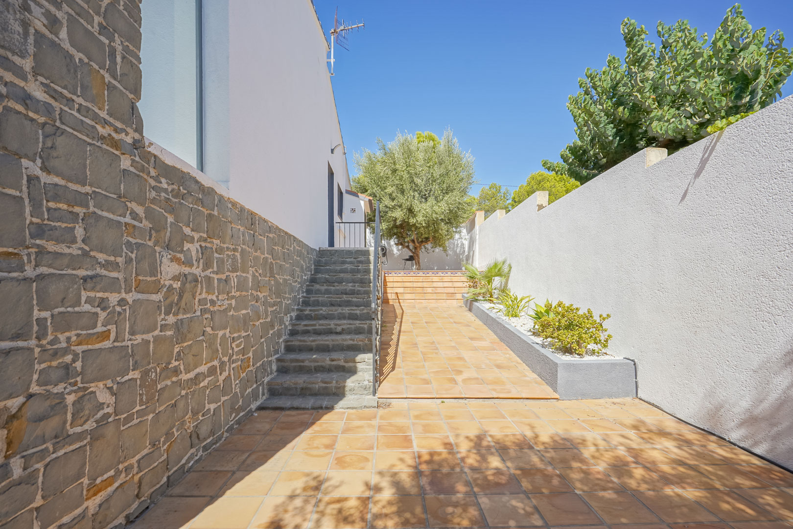 Entrada exterior con escalera de piedra natural, muros revestidos en mampostería y suelos de gres rústico junto a un jardín mediterráneo con olivos.