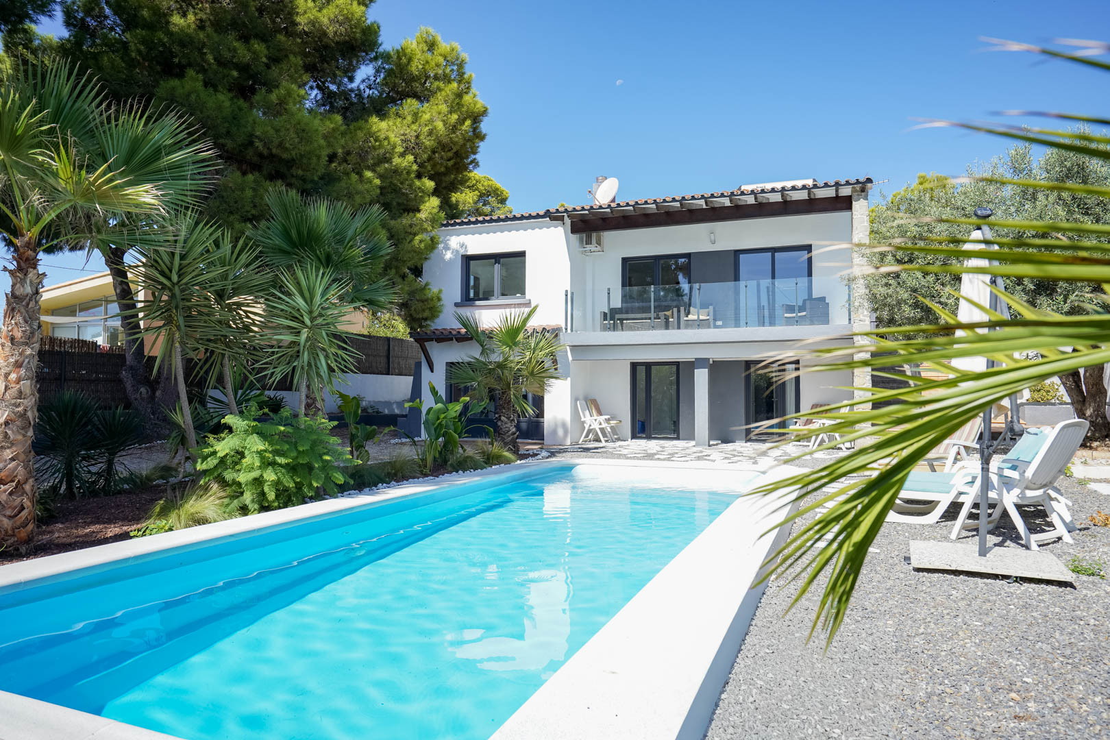 Exterior de la villa con piscina rectangular, jardín mediterráneo con palmeras y fachada blanca de dos niveles con terrazas orientadas al sur.