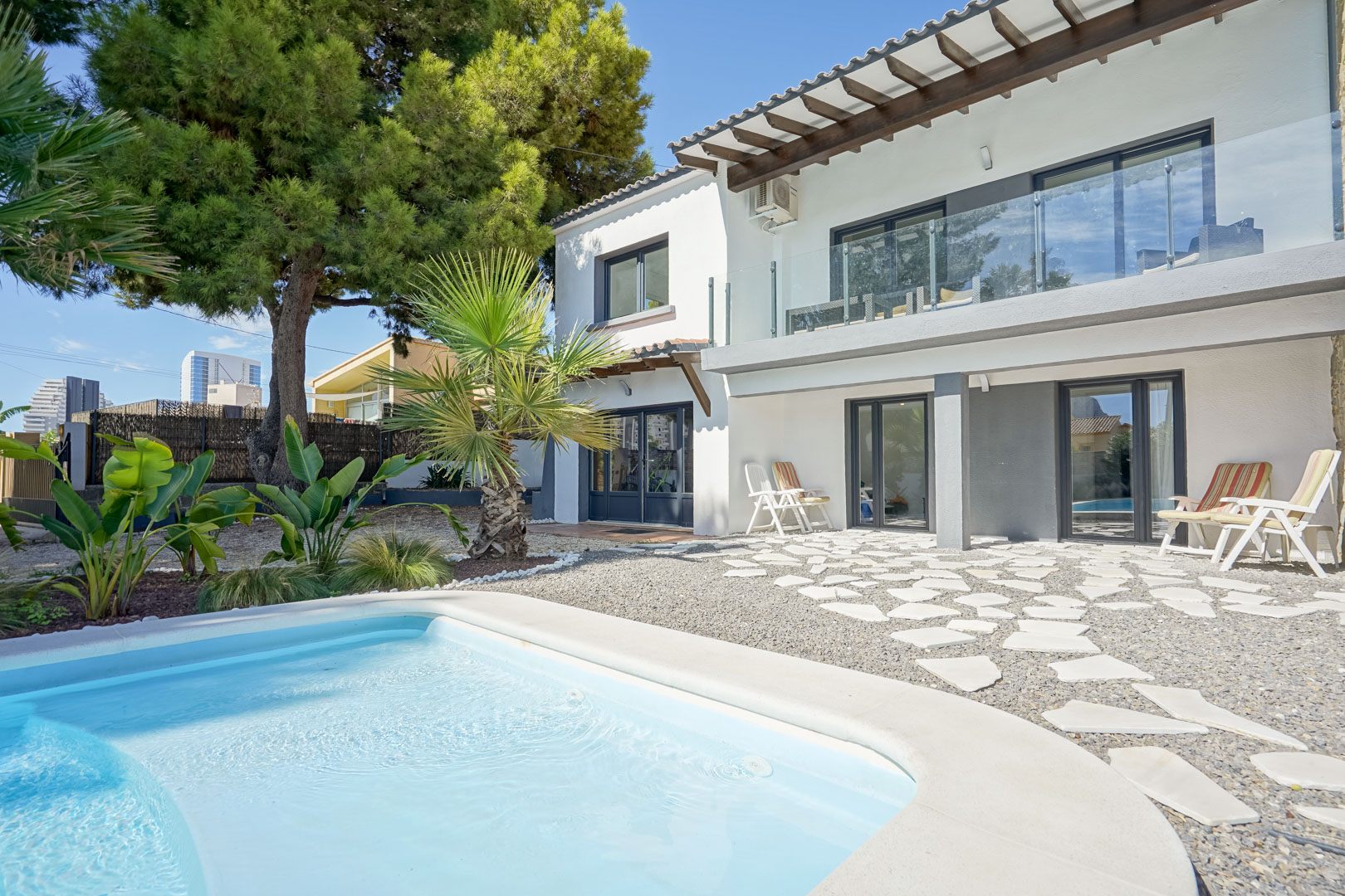 Fachada posterior de la villa con piscina, destacando la terraza superior con barandilla de cristal y vigas de madera, y zona de grava con piedras irregulares.