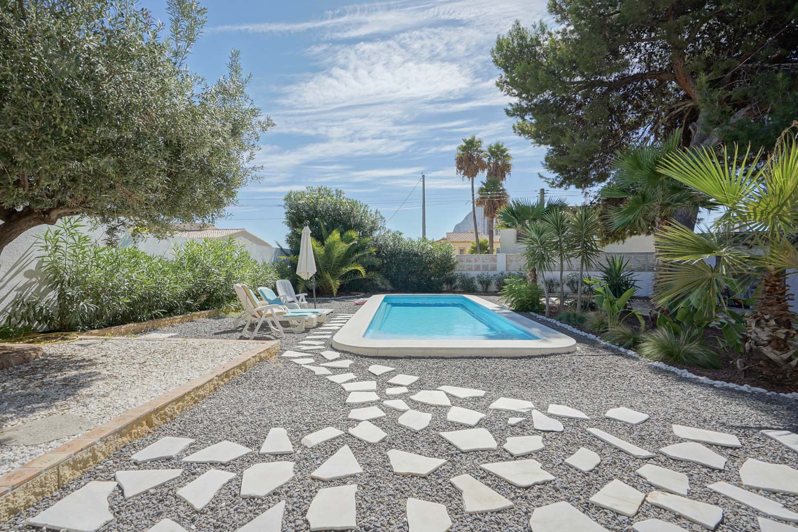 Piscina rectangular rodeada de un jardín mediterráneo con olivos y palmeras. Incluye un camino de piedras irregulares sobre grava y zona de solárium.