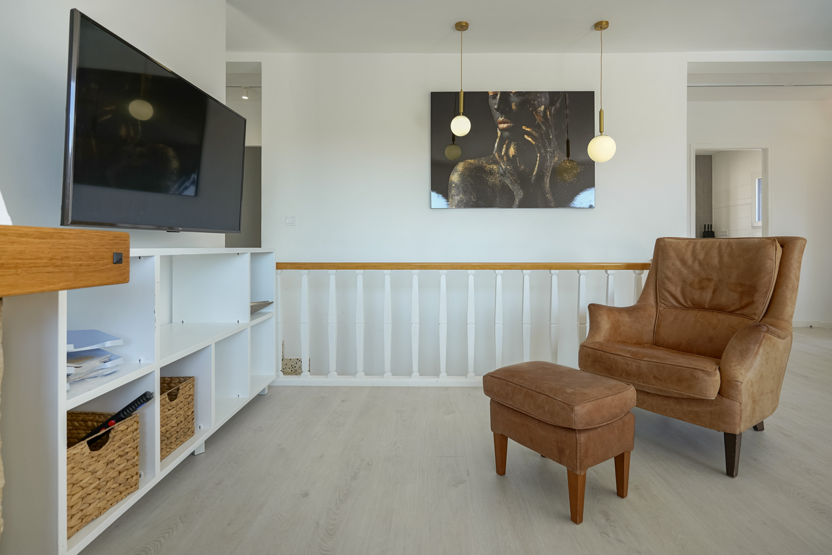 Rincón de descanso con sillón de cuero y reposapiés, estantería blanca y barandilla de madera. Incluye iluminación colgante moderna y suelos laminados claros.