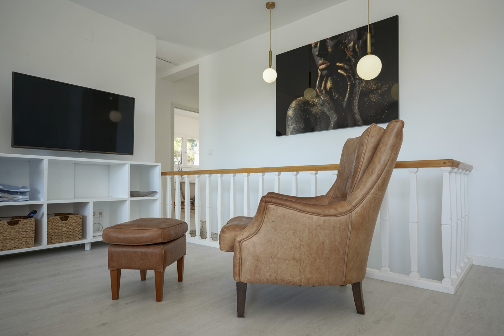 Rincón de lectura con sillón de cuero y reposapiés junto a la barandilla de la escalera. Dispone de televisión de pared, estantería blanca y lámparas colgantes.