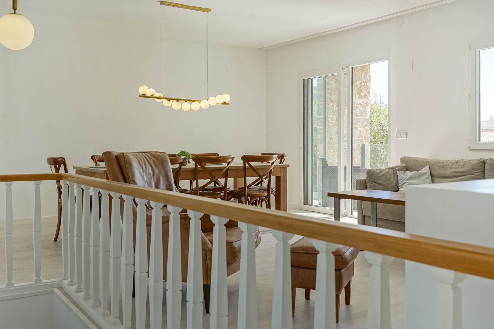 Salón-comedor luminoso con barandilla clásica, mesa de madera, iluminación de diseño y acceso directo a la terraza a través de amplios ventanales.