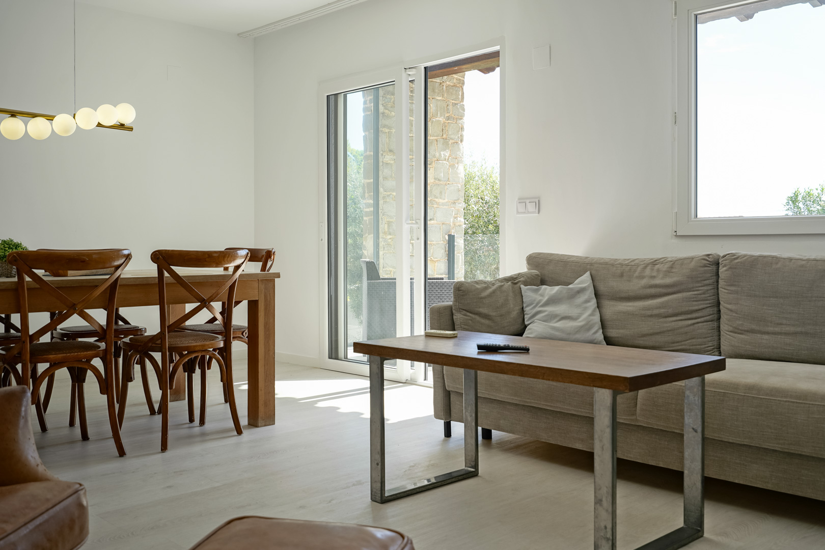 Salón-comedor luminoso con mobiliario de madera y lámpara de esferas. Los ventanales de doble acristalamiento conectan el interior con la terraza de piedra natural.