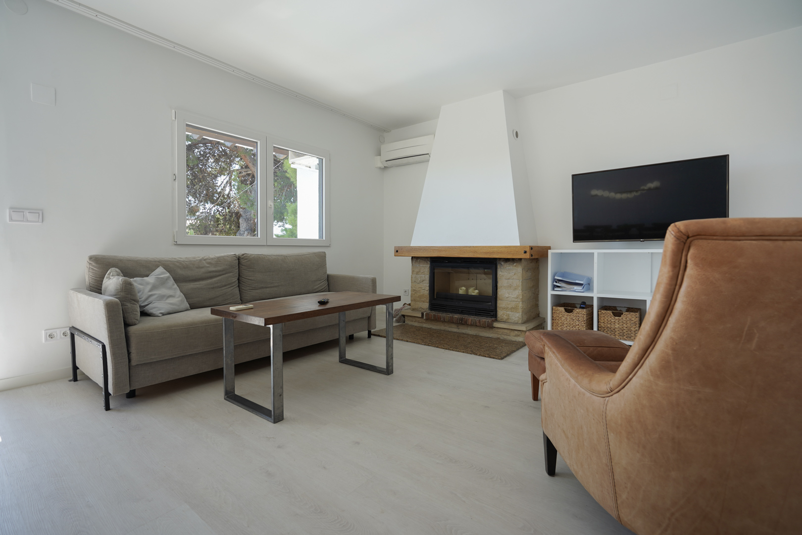 Salón moderno con chimenea de piedra y madera, sofá gris y sillón de cuero. Incluye aire acondicionado y ventanales de doble acristalamiento con vistas al exterior.