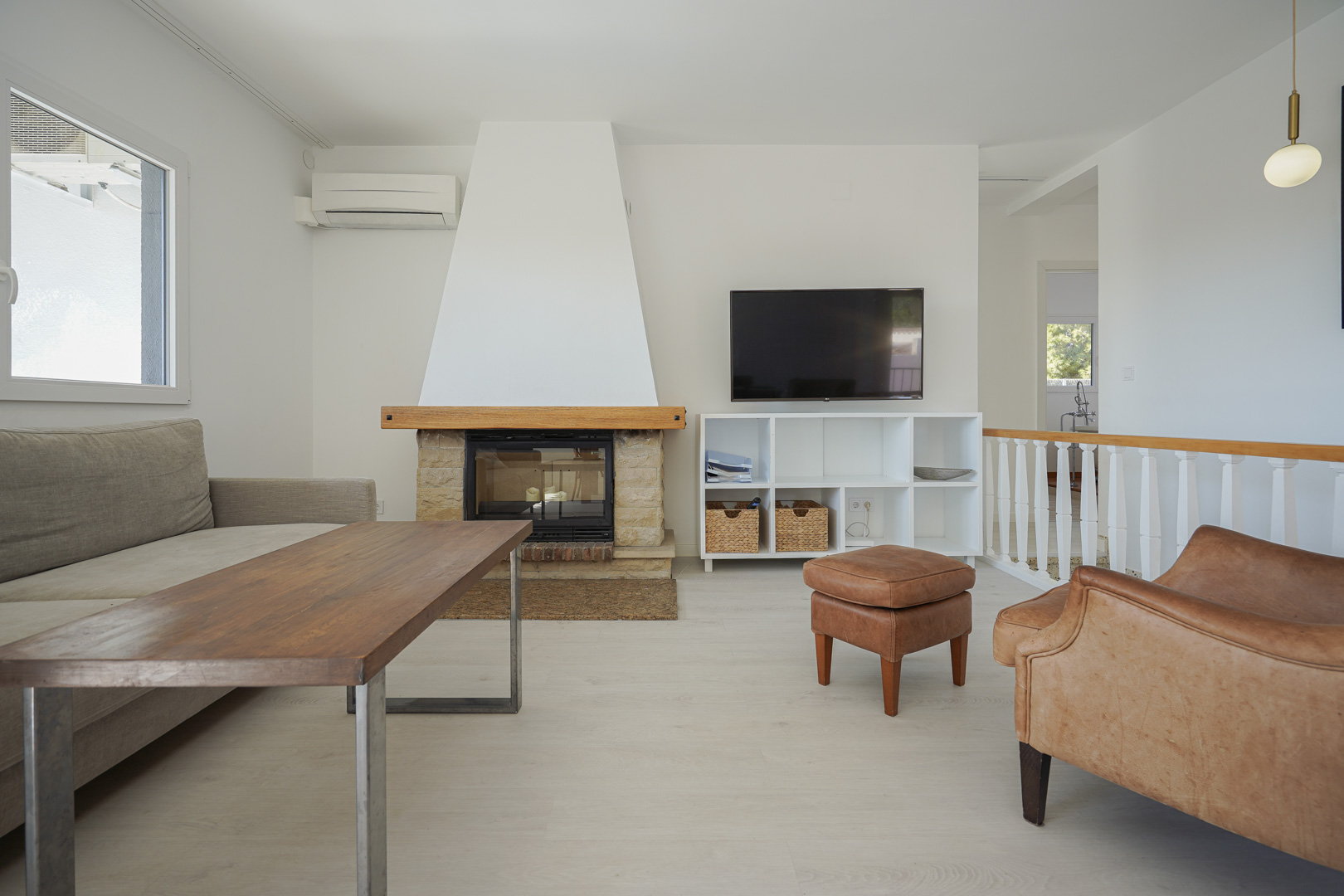 Salón moderno con chimenea de piedra y madera, suelos de tarima clara, aire acondicionado y acceso a la escalera con barandilla blanca.