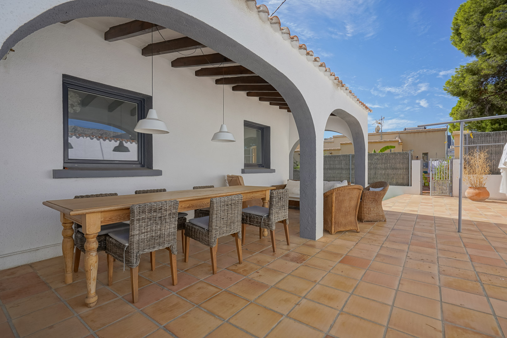 Terraza cubierta con arcos blancos, vigas de madera vista y suelo de terracota. Dispone de una zona de comedor con mesa de madera y sillas de mimbre.
