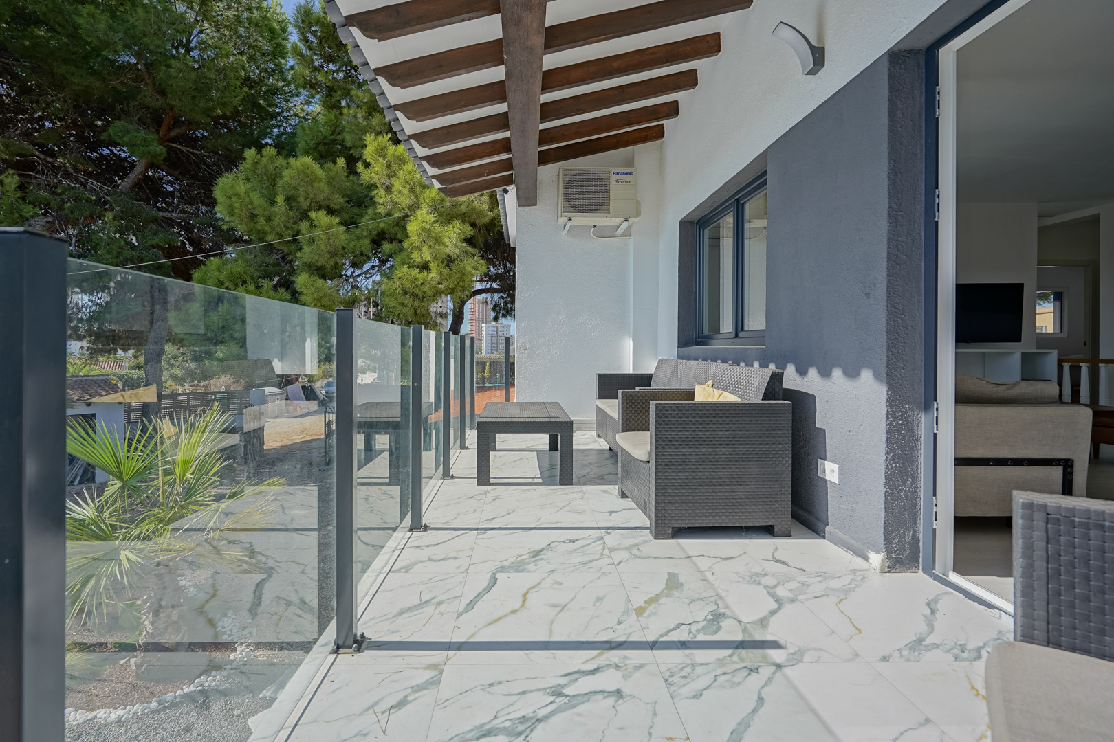Terraza cubierta con suelo de mármol veteado, barandilla de cristal y techo de vigas de madera. Incluye mobiliario de ratán y vistas al entorno arbolado de la propiedad.