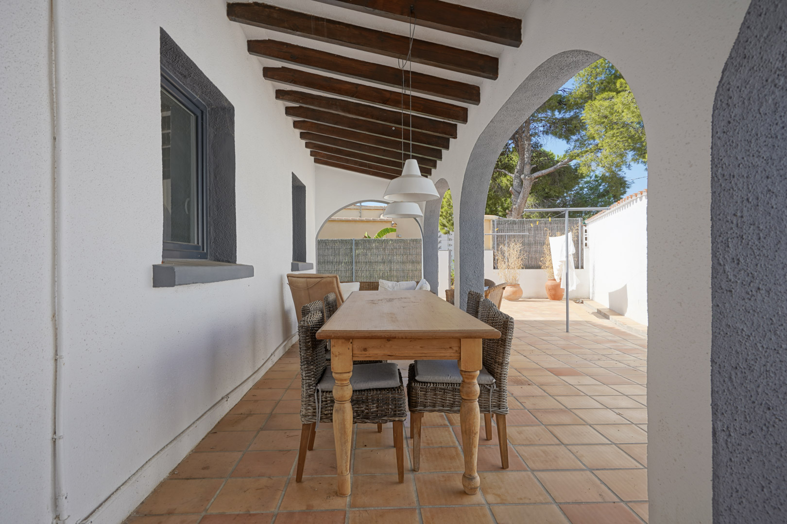 Terraza cubierta con vigas de madera vista, arcos mediterráneos y zona de comedor exterior con mesa de madera y sillas de mimbre sobre suelo de terracota.