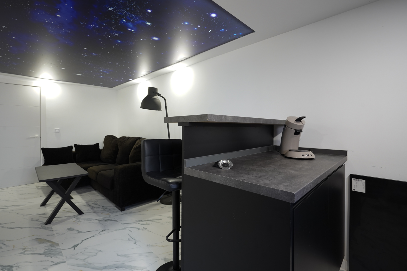 Zona de estar con barra de café, sofá oscuro y suelo de gres efecto mármol, destacando un techo retroiluminado con diseño de cielo estrellado.