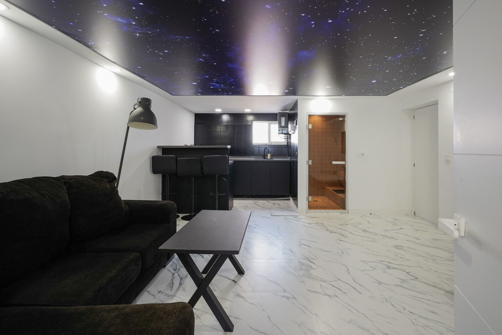 Zona de estar con cocina americana, suelos de gres porcelánico efecto mármol, techo tensado con diseño de galaxia y acceso a una sauna integrada.