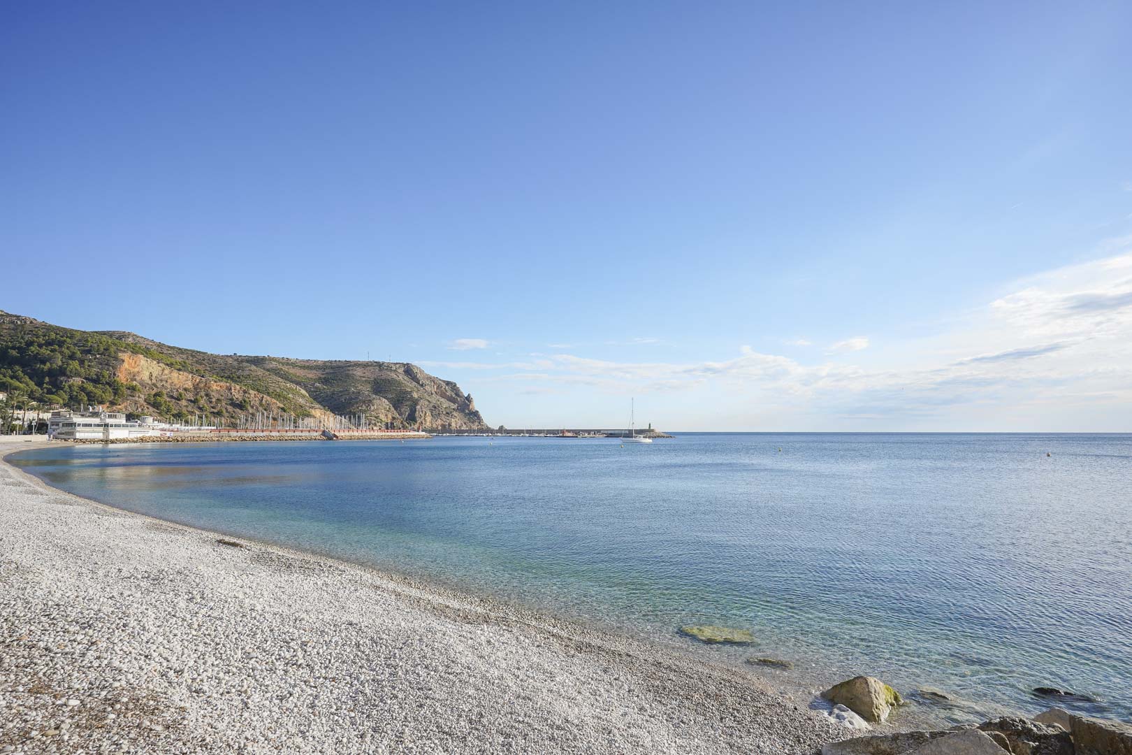 Vista panorámica de la playa de grava en Jávea, mostrando aguas cristalinas del Mediterráneo y el puerto bajo el acantilado del Cabo de San Antonio.