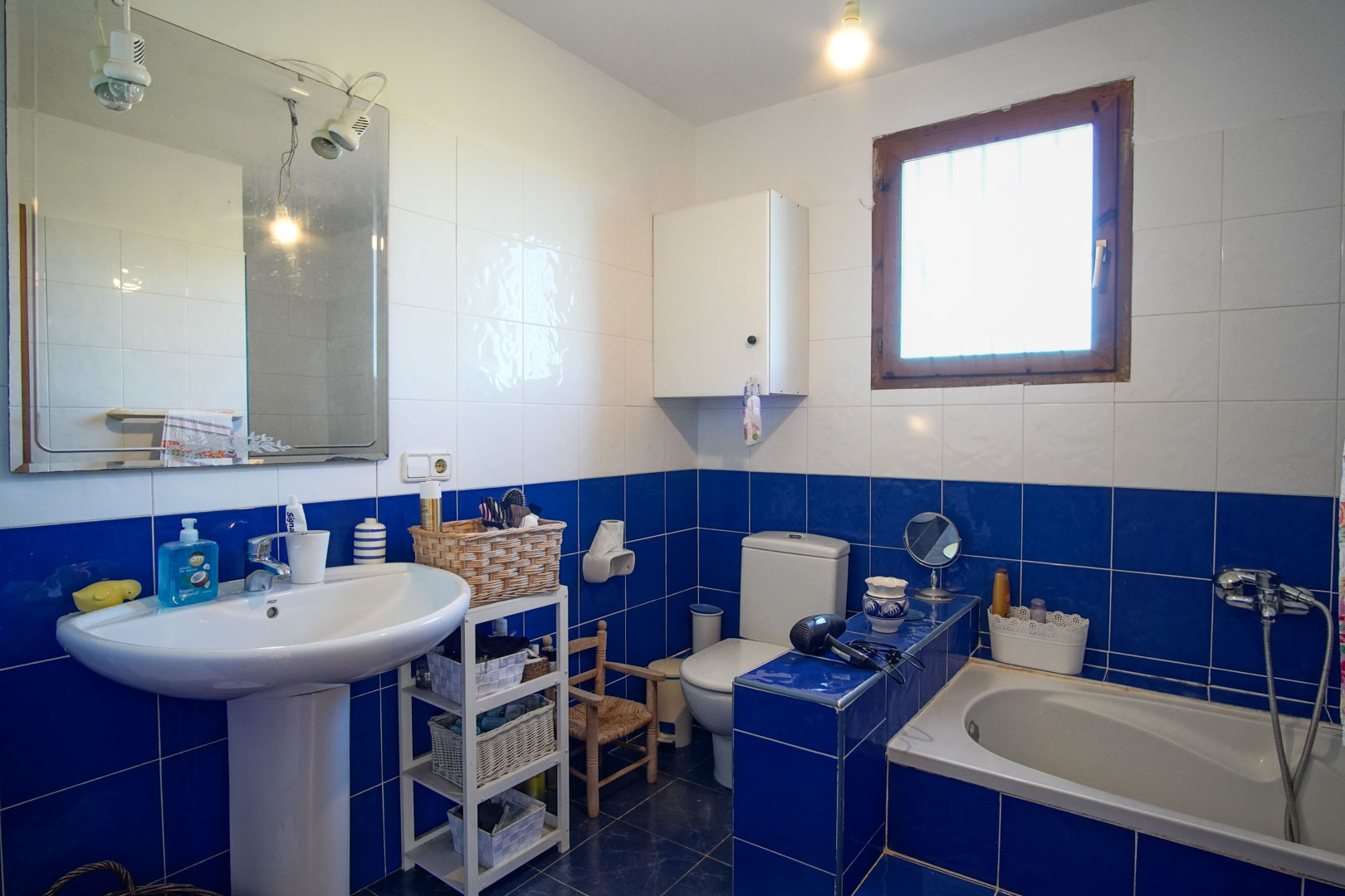 Baño con azulejos en azul y blanco, equipado con lavabo de pedestal, bañera integrada y ventana con marco de madera que aporta luz natural.