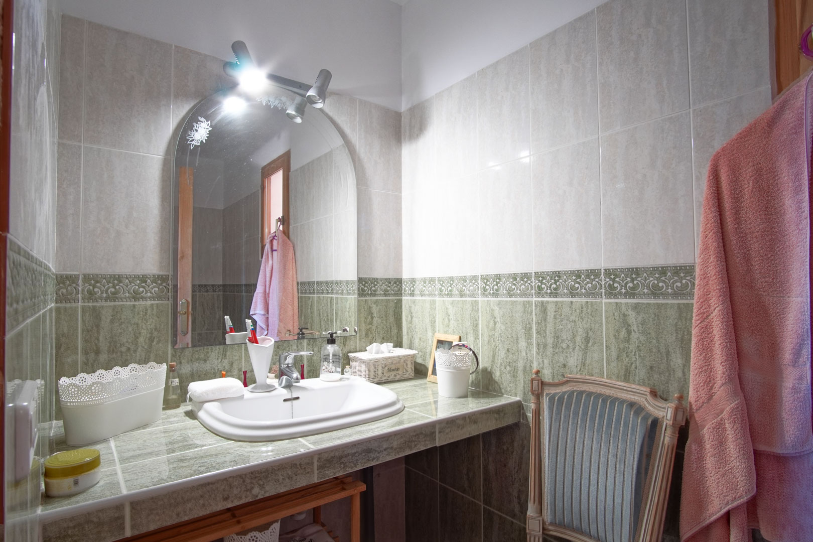 Cuarto de baño con encimera alicatada en tonos verdes, lavabo encastrado, espejo de arco con focos superiores y paredes con cenefa decorativa.