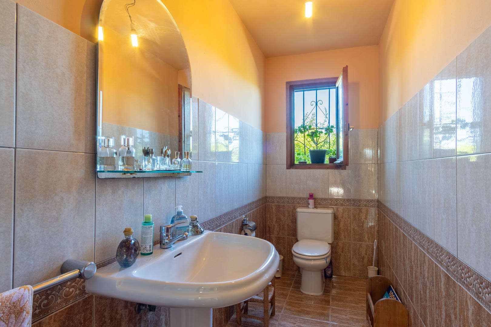 Cuarto de baño con paredes alicatadas en tonos tierra, lavabo de pedestal, espejo de arco y ventana de madera con vistas al exterior.