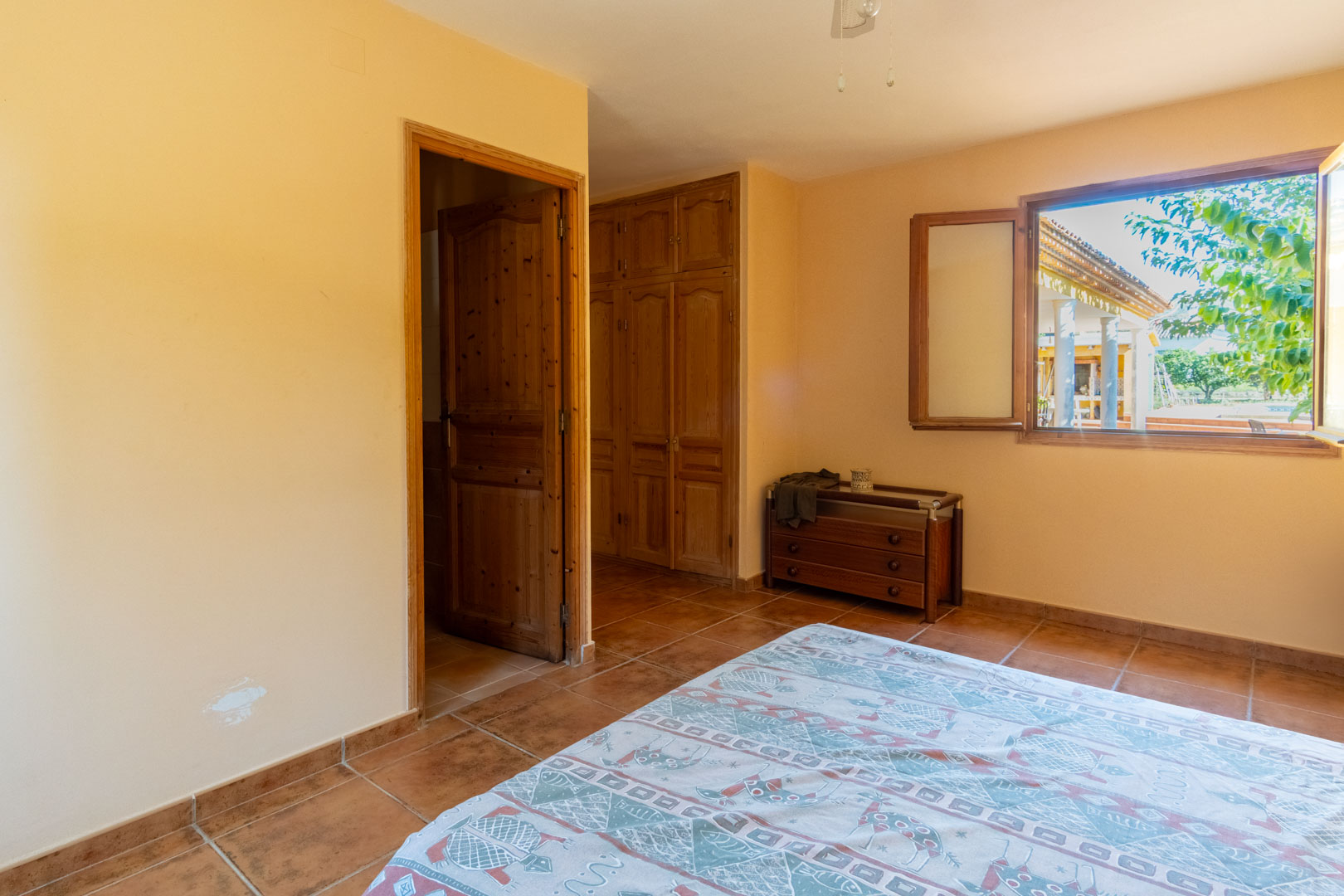 Dormitorio con suelos de gres rústico, armarios empotrados de madera y ventana con vistas al porche exterior y zonas verdes.