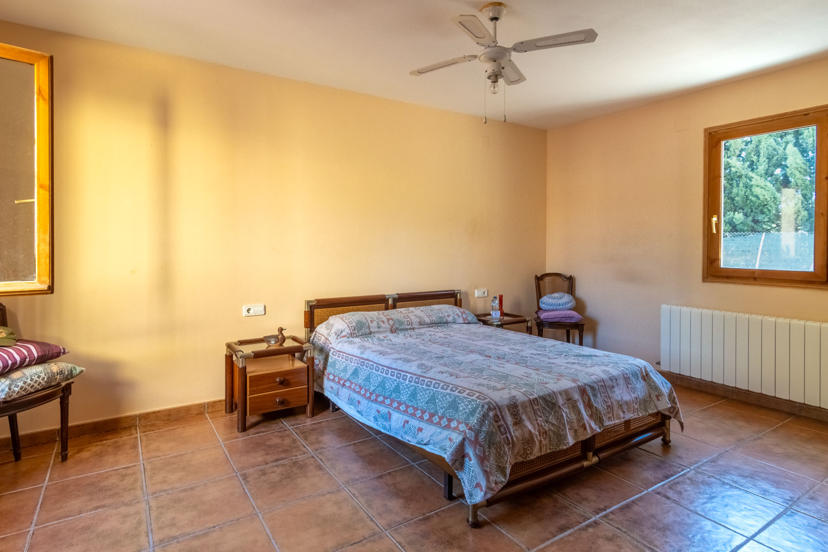 Dormitorio con suelos de terracota, paredes en tonos cálidos y ventilador de techo. Incluye ventanas de madera, radiador y mobiliario de estilo rústico con mesitas de noche a juego.