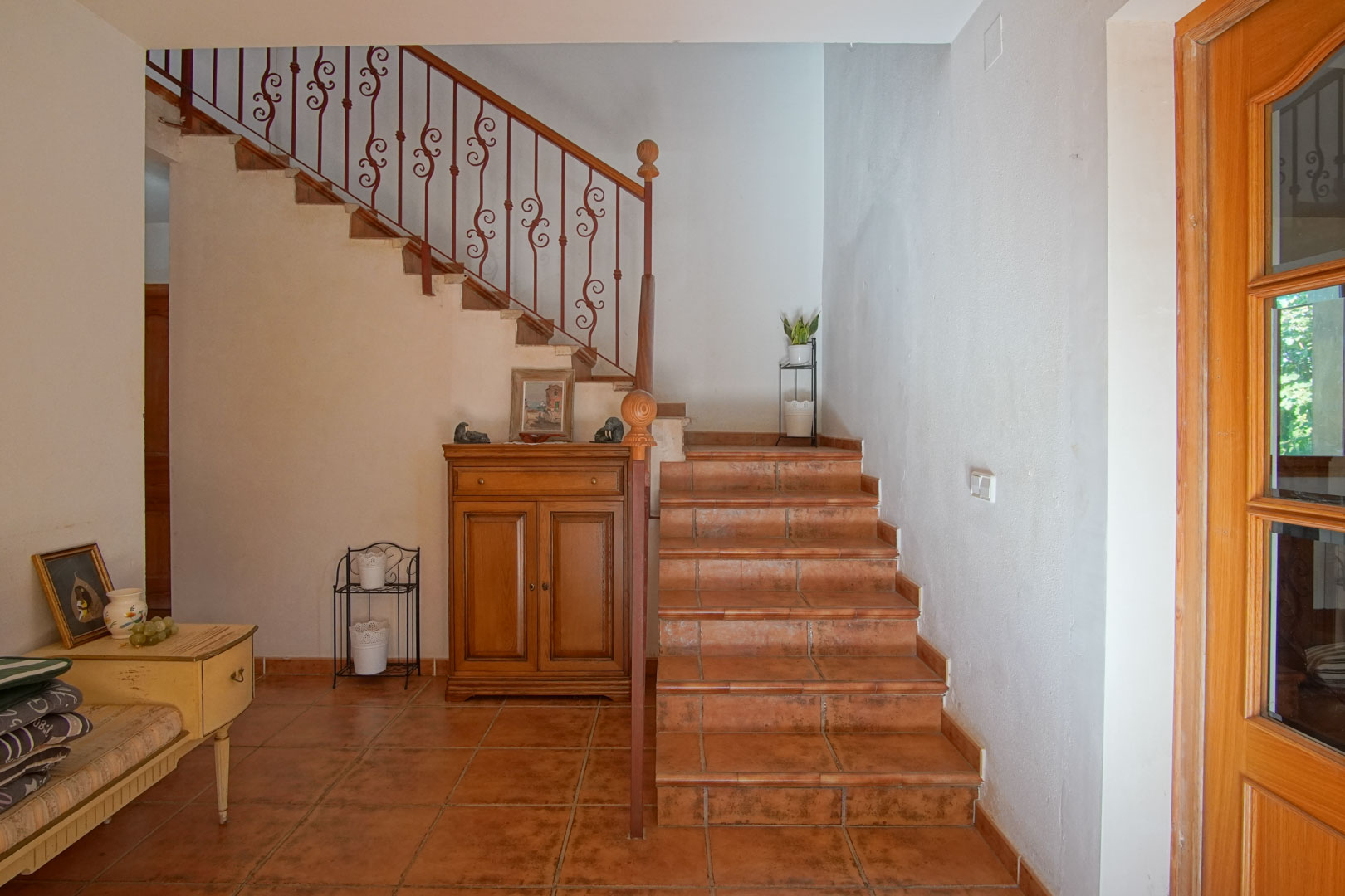 Escalera interior con peldaños de gres rústico y barandilla de hierro forjado ornamentada. El recibidor cuenta con suelos de terracota y mobiliario de madera de estilo clásico.