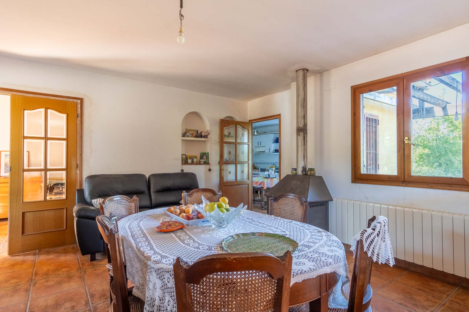 Salón-comedor con suelos de terracota, mesa de madera con sillas de rejilla y una chimenea metálica. La estancia destaca por su carpintería de madera y la entrada de luz natural.