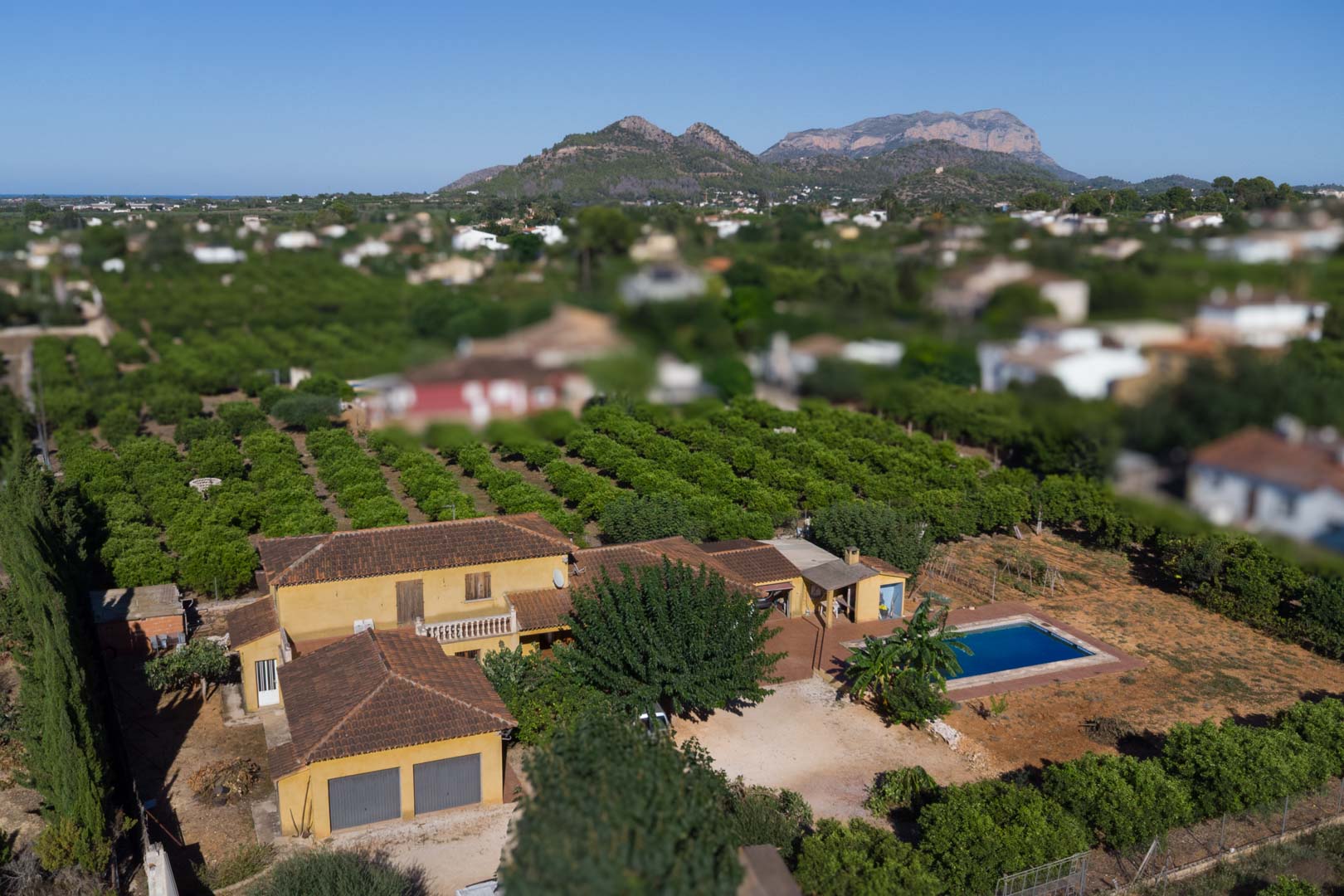 Vista aérea de la propiedad que muestra la vivienda de estilo rústico, su piscina privada y el extenso huerto de cítricos con la montaña de Segaria al fondo.