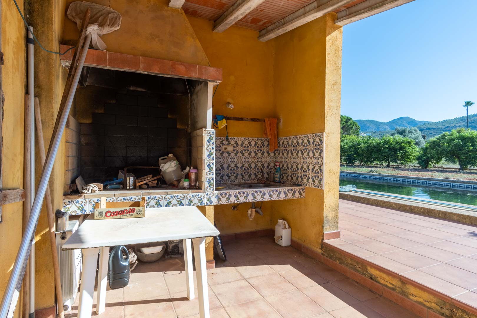 Zona de paellero exterior con barbacoa de obra, fregadero con azulejos decorativos y vistas directas a la piscina, el huerto y las montañas de Segaria.