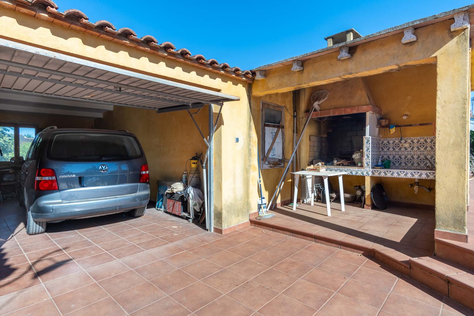 Zona exterior funcional que incluye un parking cubierto para vehículos y un paellero tradicional con barbacoa de obra y encimera alicatada.