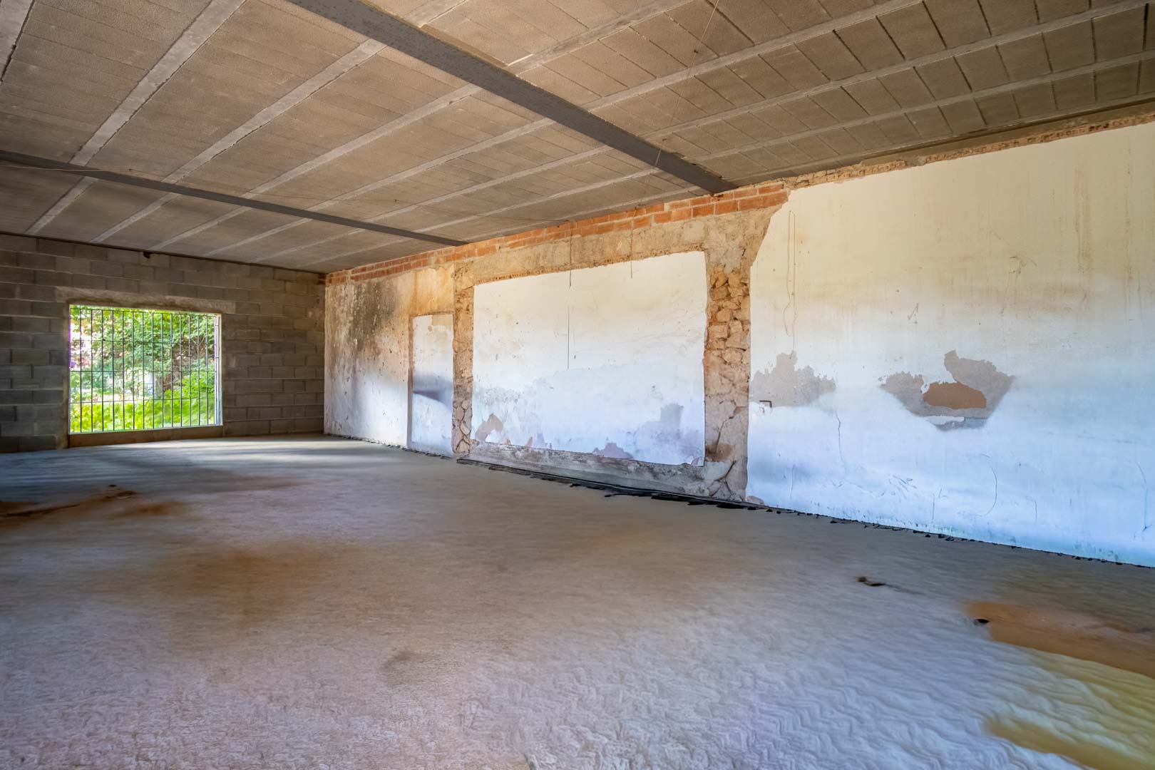 Espacio interior diáfano en fase de reforma con suelos de hormigón, techos de viguetas vistas y muros que combinan piedra original con ladrillo.