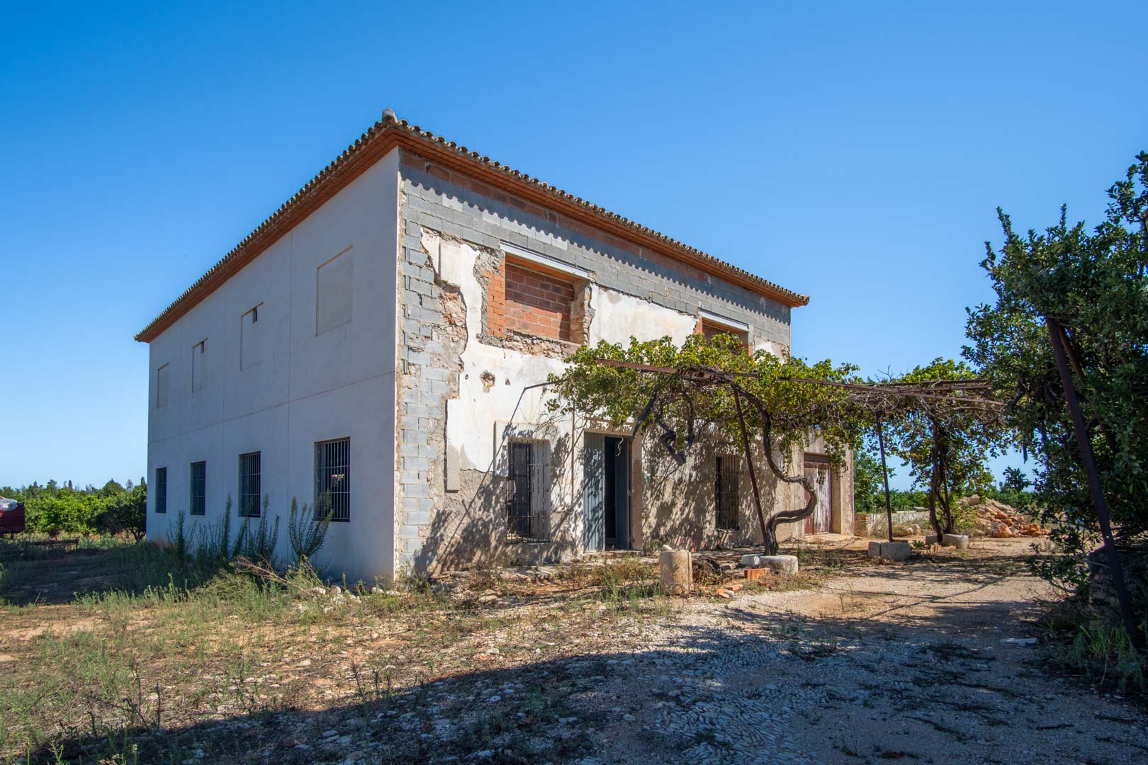 Fachada de casa de campo en proceso de reforma con estructura consolidada, porche de parra natural y amplias posibilidades de personalización en parcela llana.