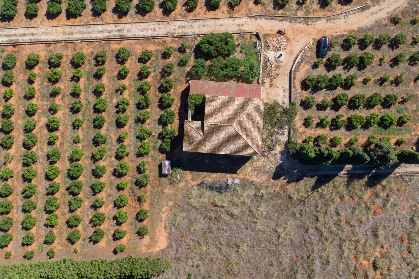 Vista aérea cenital de la casa de campo con tejado de teja tradicional, rodeada de extensas hileras de cultivos en una parcela plana y soleada en Dénia.