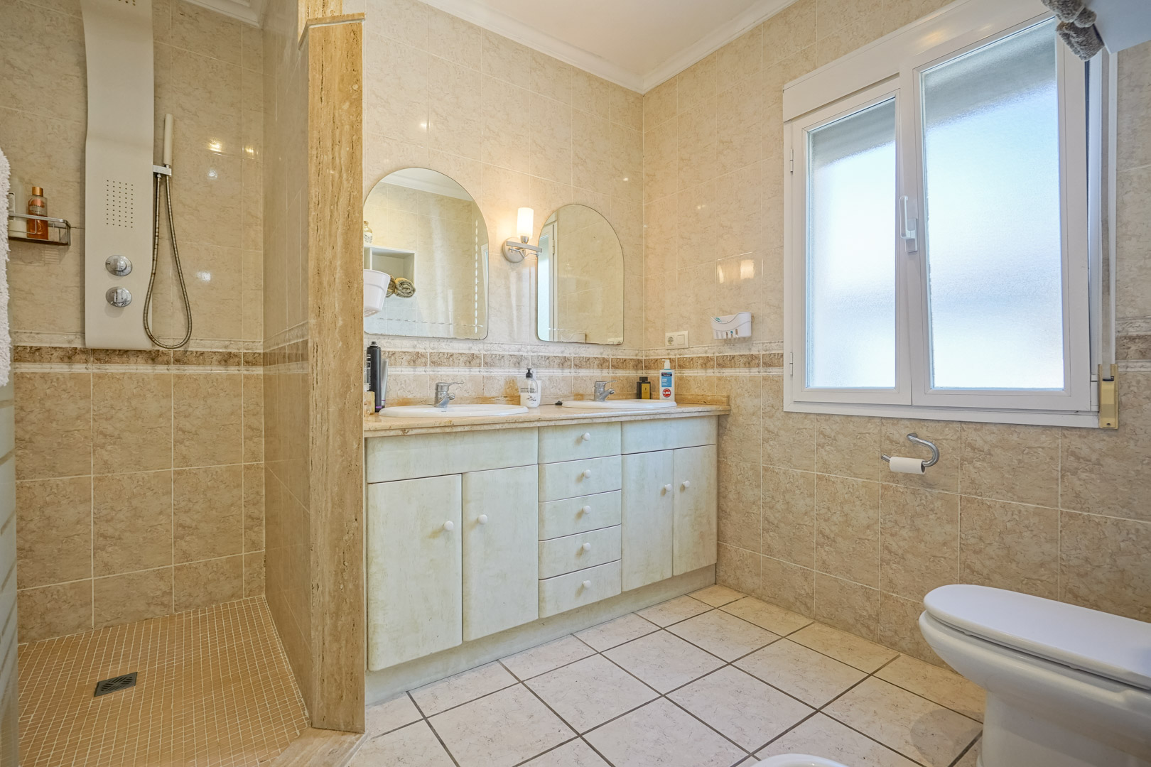 Cuarto de baño con ducha a ras de suelo, tocador con doble lavabo y azulejos color crema.