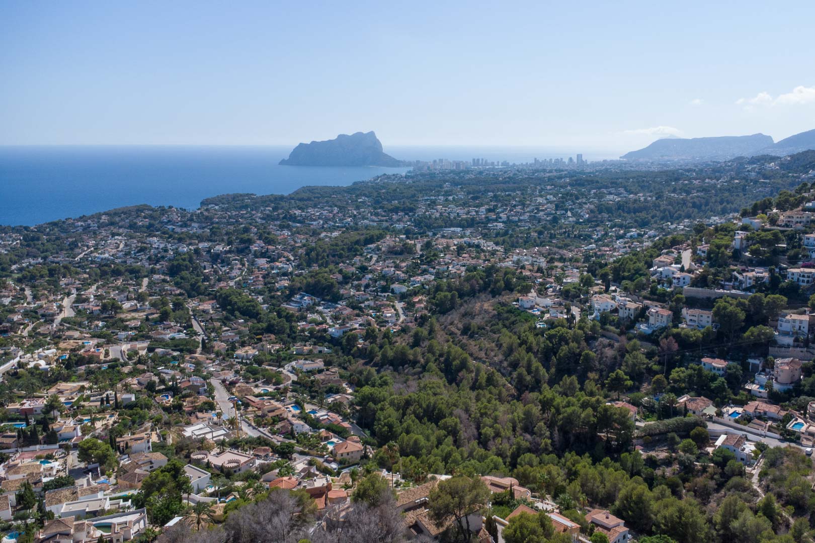 Vista aérea panorámica de la costa de Moraira y Calpe, destacando el Peñón de Ifach sobre el mar Mediterráneo y las colinas residenciales arboladas.