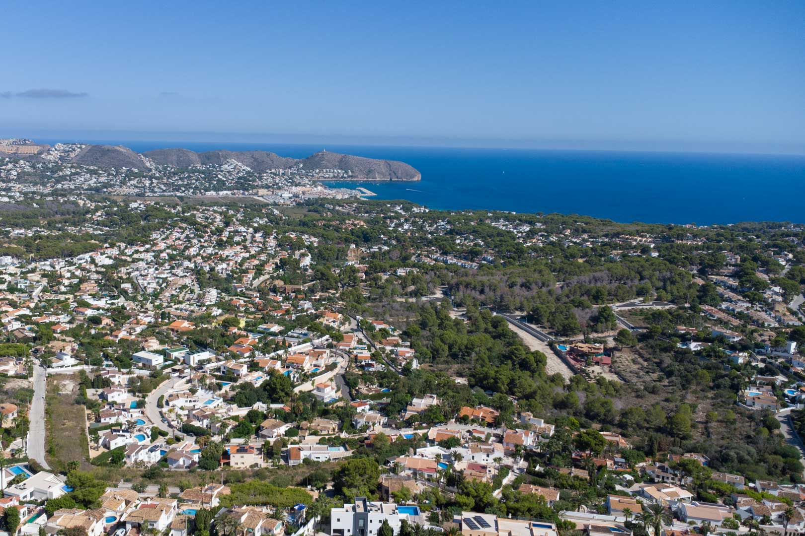 Vista aérea panorámica de Moraira que muestra la costa mediterránea, el puerto y el Cap d'Or, rodeados de zonas residenciales integradas en el paisaje de pinos.