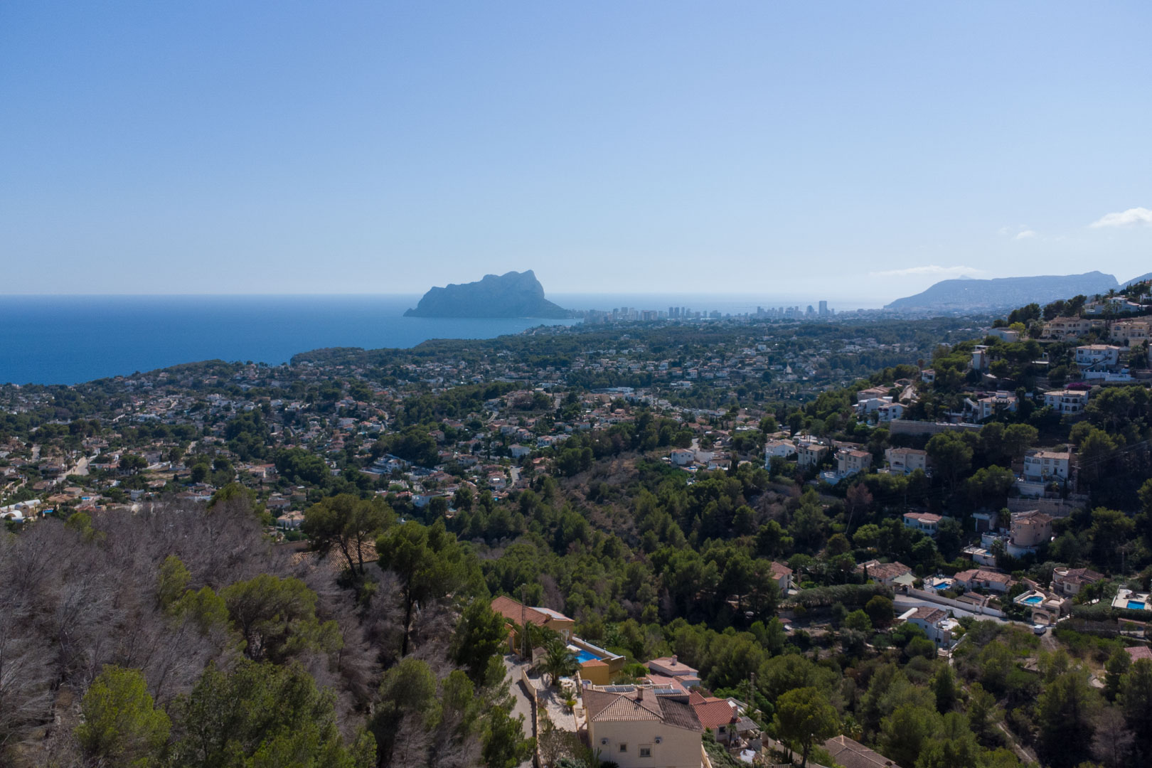 Vista panorámica desde la colina hacia el Peñón de Ifach y el mar Mediterráneo, rodeada de la exuberante vegetación y villas de la Costa Blanca.