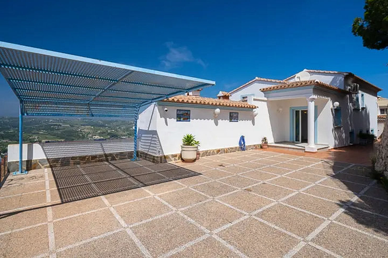 Amplio aparcamiento exterior con suelo de baldosas y una estructura de sombra metálica en azul, ofreciendo vistas despejadas a las montañas de Moraira.
