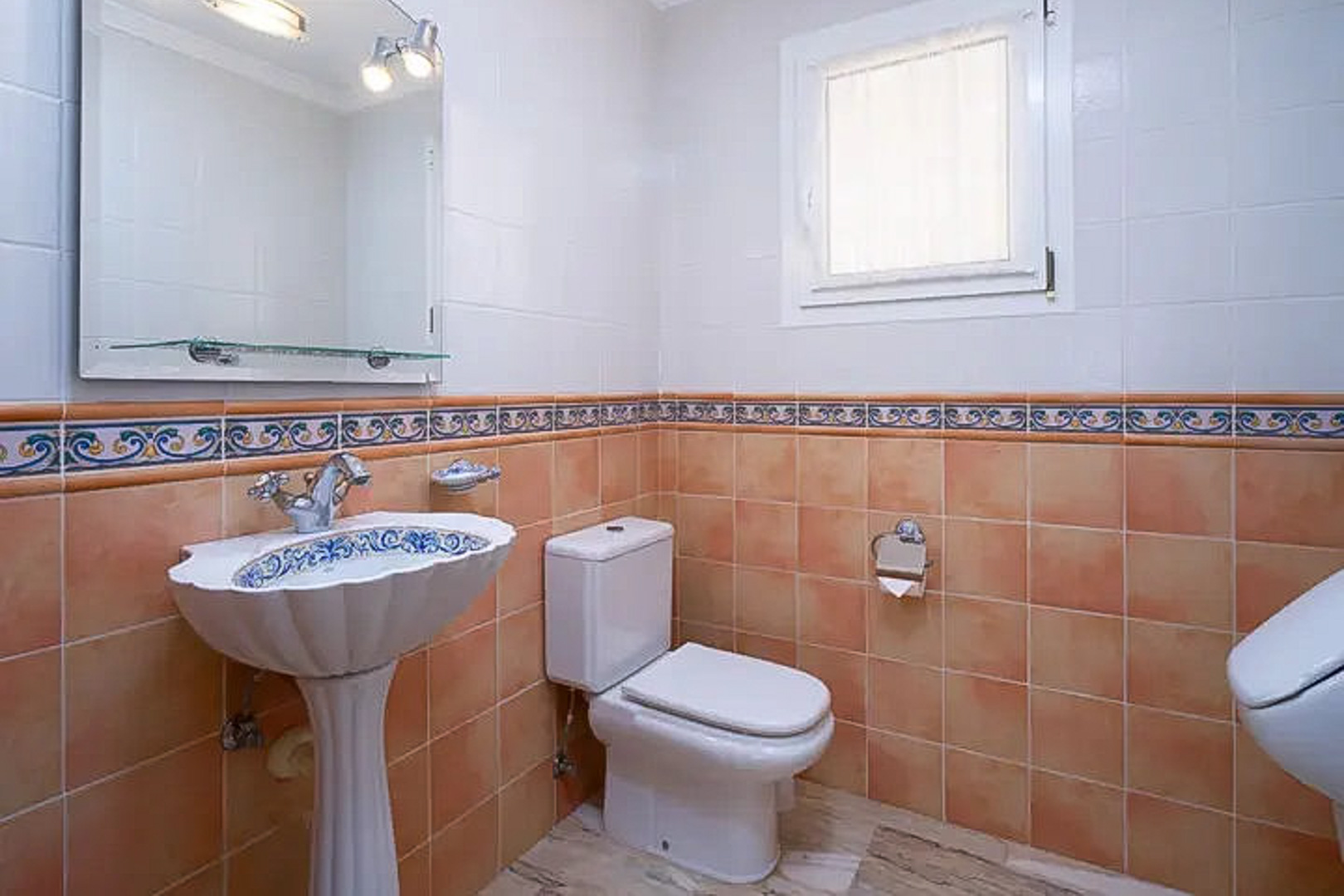 Baño de cortesía con lavabo de pedestal decorado, azulejos en tonos terracota y blanco con cenefa ornamental, espejo con iluminación y ventana de ventilación.