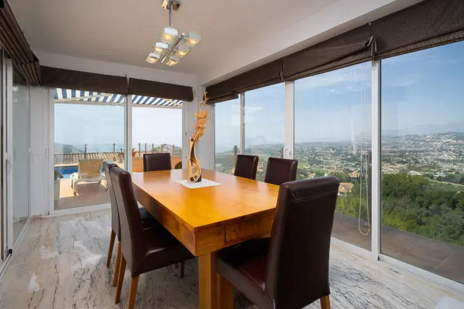 Comedor luminoso con mesa de madera maciza y sillas de cuero, rodeado de ventanales con vistas panorámicas al mar y acceso directo a la terraza de la piscina.