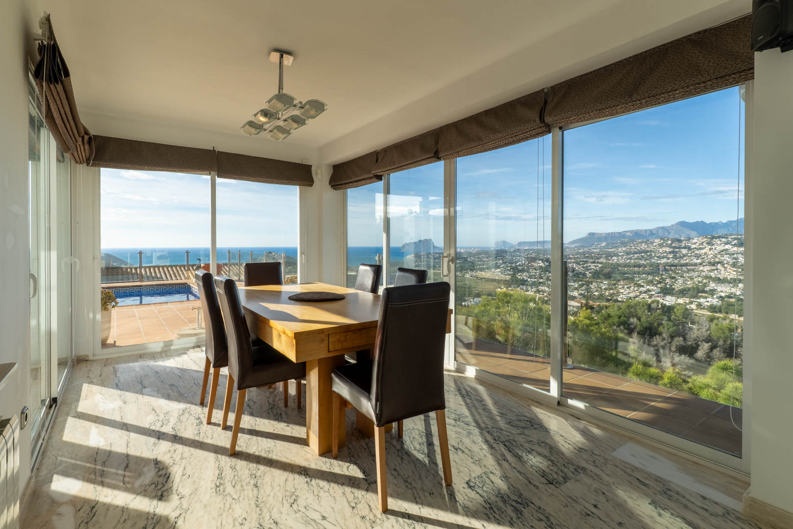 Comedor luminoso con suelo de mármol y mesa de madera maciza. Amplios ventanales ofrecen vistas panorámicas al mar Mediterráneo, el Peñón de Ifach y las montañas de Moraira.