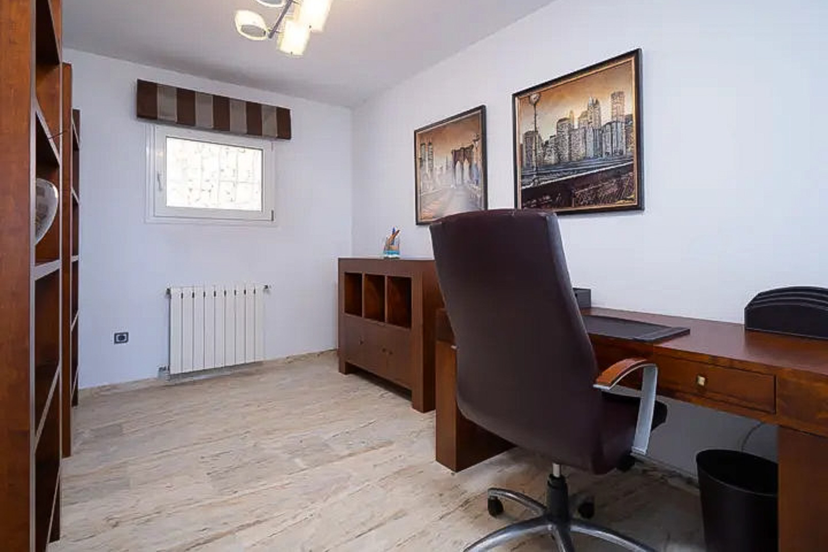 Despacho luminoso con mobiliario de madera, escritorio funcional y silla de cuero. Incluye estanterías, suelo de piedra clara y cuadros decorativos de paisajes urbanos.