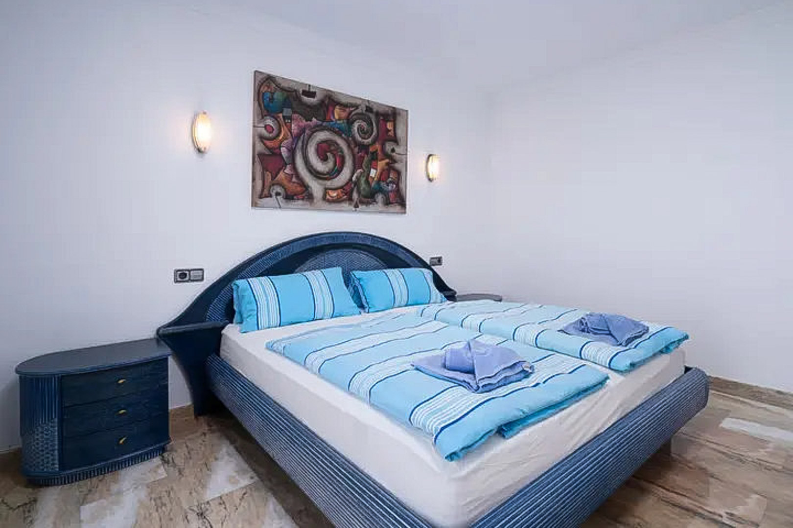 Dormitorio con cama doble, cabecero de mimbre azul y mesita a juego. Incluye suelo de mármol pulido, apliques de pared y un cuadro abstracto decorativo.