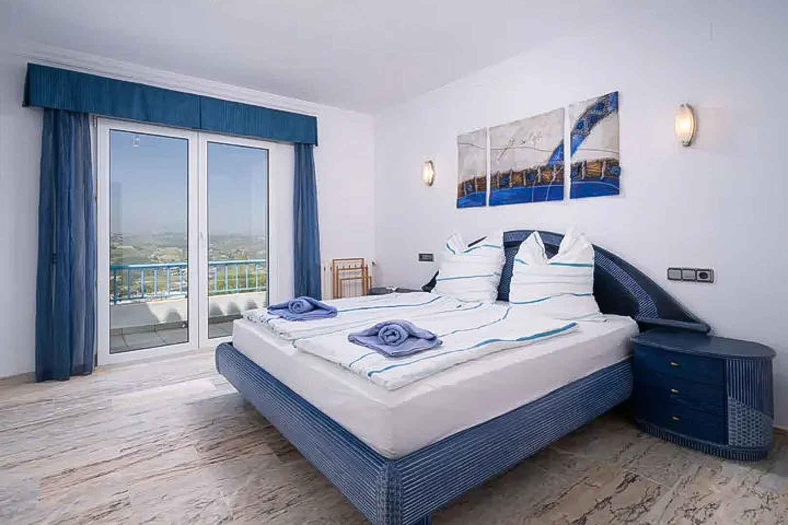 Dormitorio con suelo de mármol, cama doble en tonos azules y acceso directo a una terraza con vistas panorámicas al entorno de Moraira.