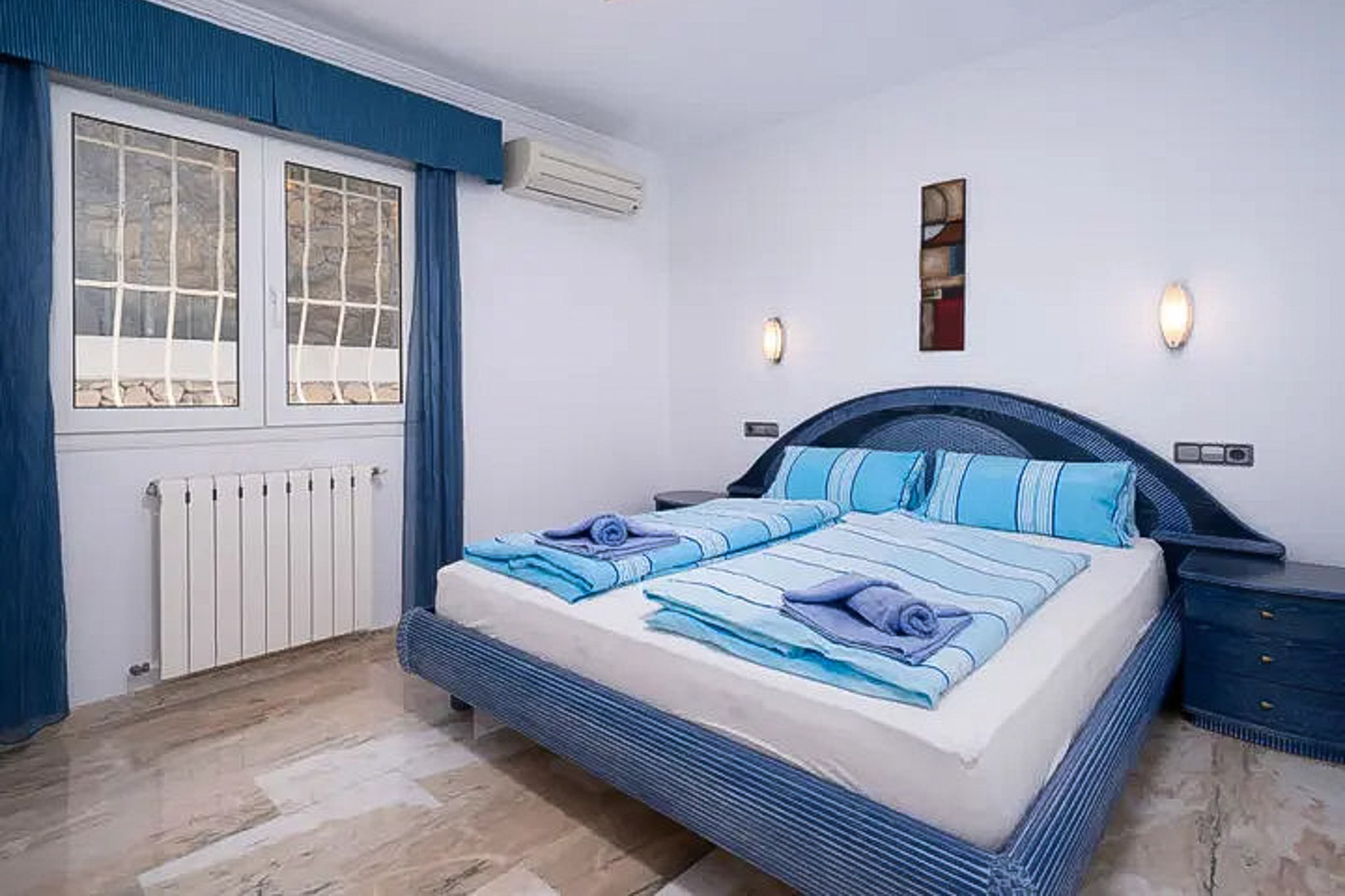 Dormitorio con suelos de mármol pulido, cama doble en tonos azules, aire acondicionado, radiador y ventana exterior con rejas de seguridad.