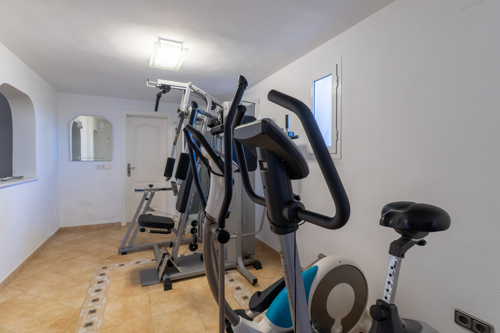 Gimnasio privado equipado con máquina de musculación, elíptica y bicicleta estática sobre suelo cerámico con cenefas decorativas.