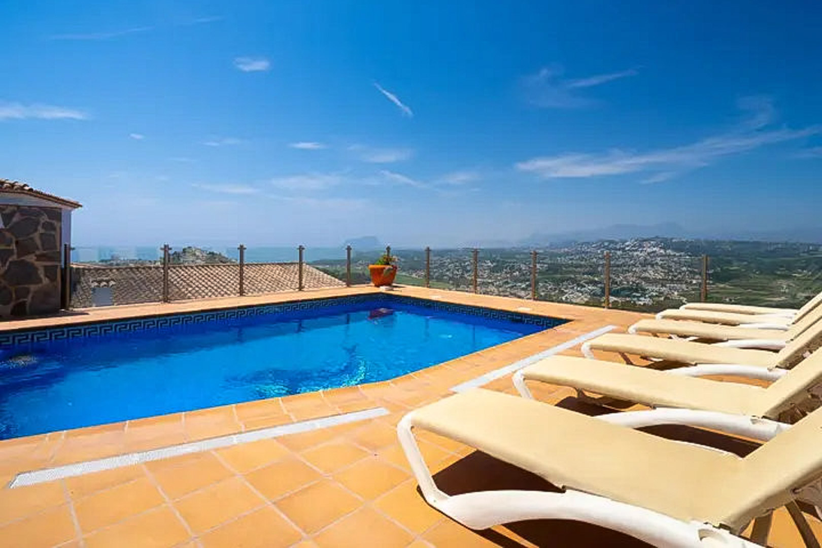 Piscina exterior con borde de mosaico griego, terraza de terracota con tumbonas blancas y barandilla de cristal con vistas panorámicas al mar y Moraira.