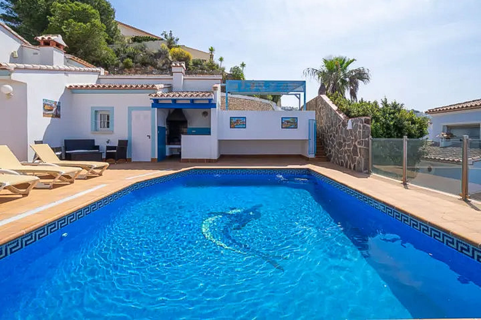 Piscina exterior con mosaico de delfín, rodeada de una terraza con tumbonas, zona de barbacoa integrada y acabados en blanco y azul de estilo mediterráneo.