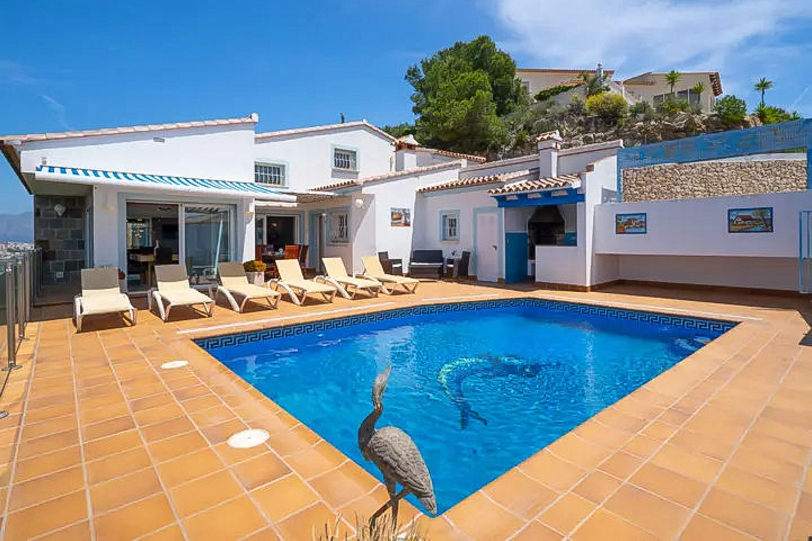 Piscina exterior con mosaico de delfín, rodeada de una terraza de gres y tumbonas bajo la fachada blanca con detalles azules y zona de barbacoa.