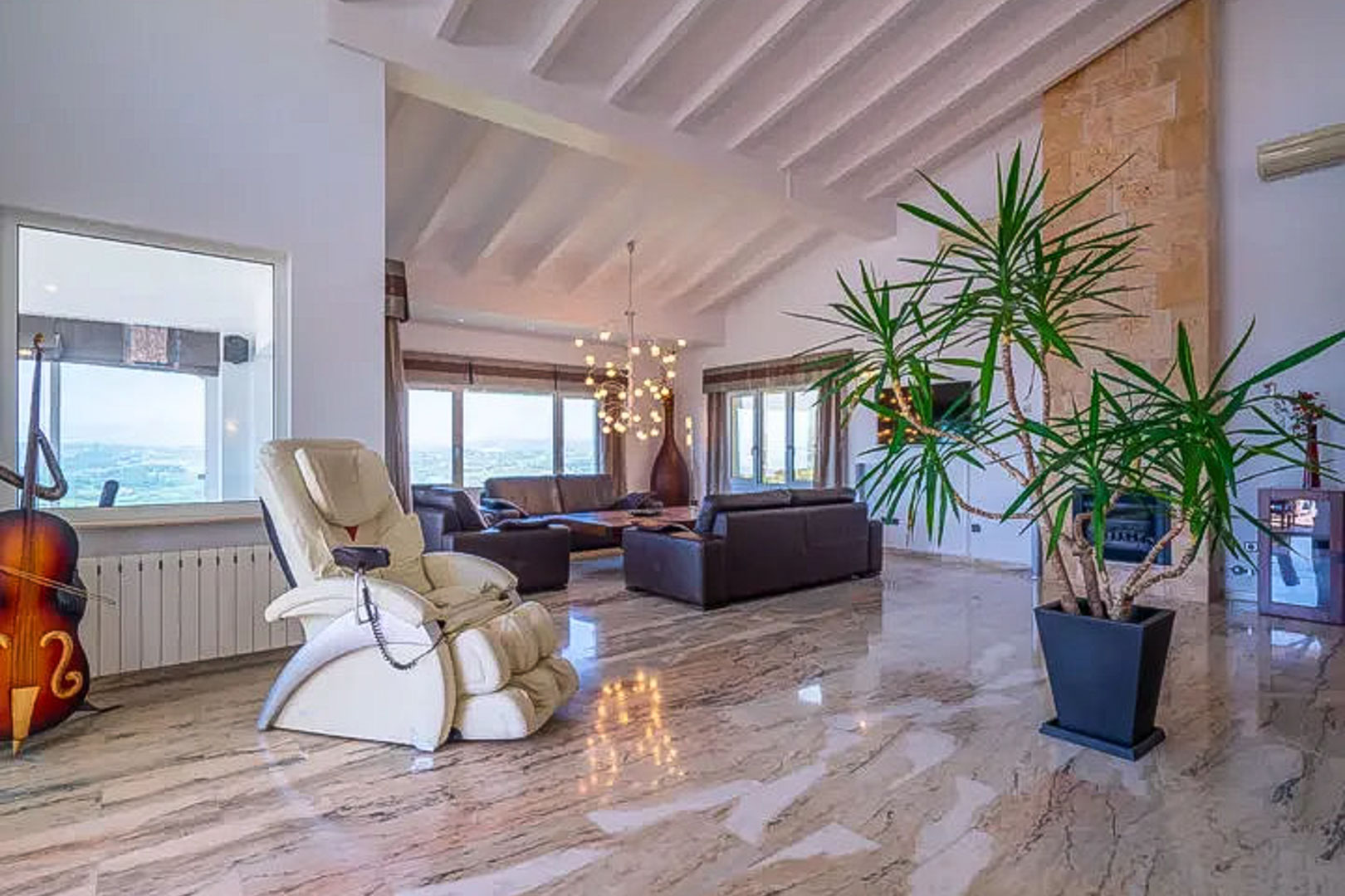 Salón amplio con techos de vigas blancas, suelo de mármol pulido y chimenea de piedra. Incluye zona de descanso con sofás de cuero y vistas panorámicas al exterior.