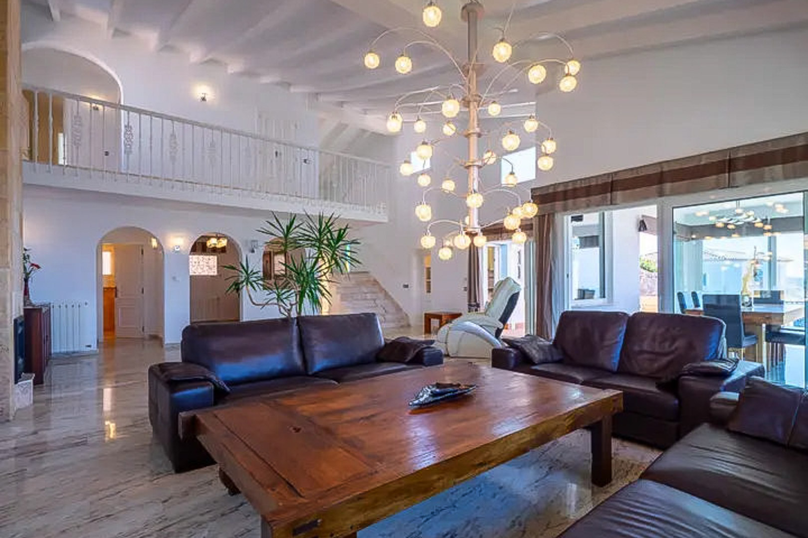 Salón de doble altura con sofás de cuero oscuro, mesa de centro de madera maciza y lámpara de diseño. Destacan los suelos de mármol y la galería superior con barandilla blanca.