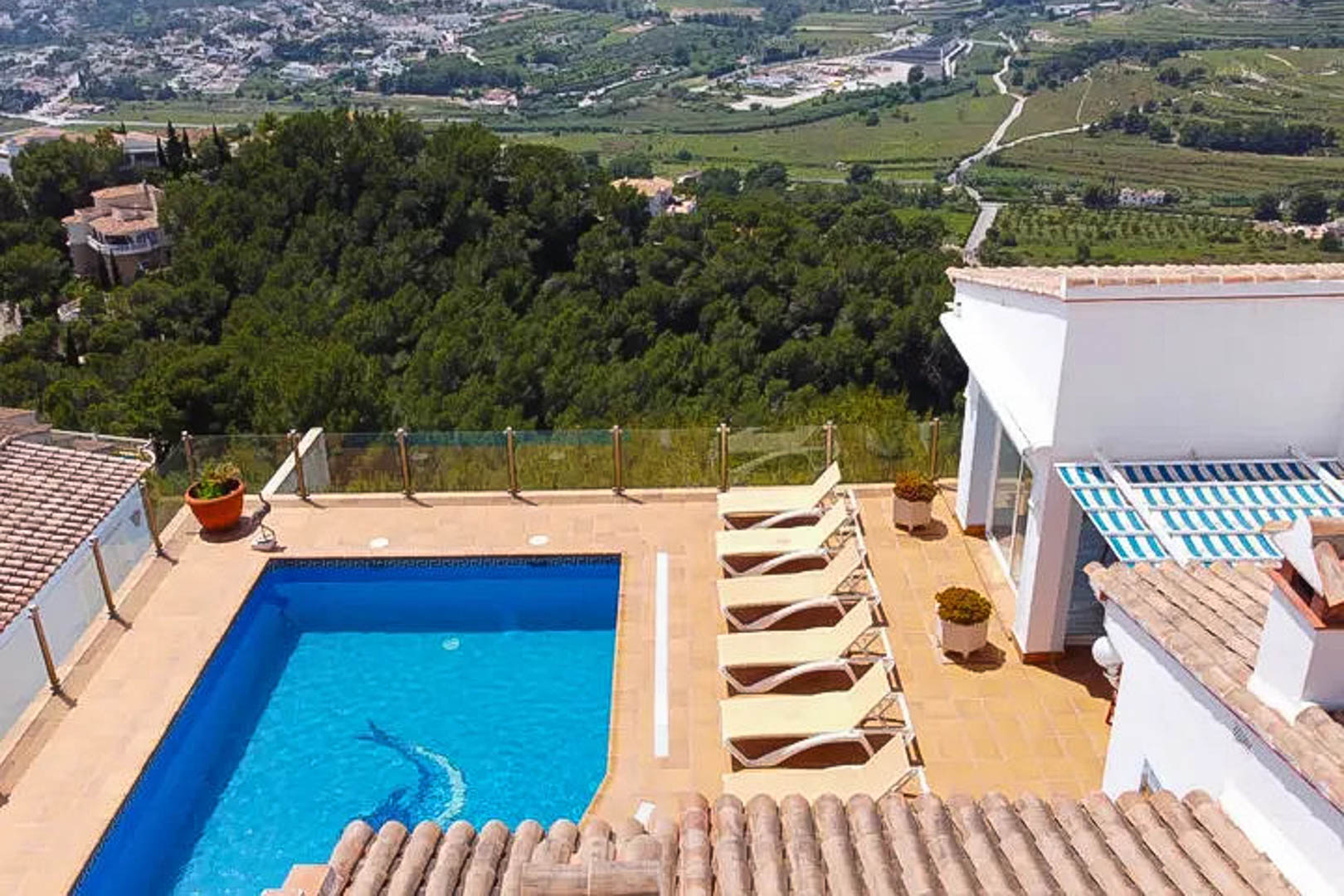 Terraza con piscina rectangular y mosaico de delfín, equipada con tumbonas y barandilla de cristal con vistas panorámicas al valle y las montañas de Moraira.