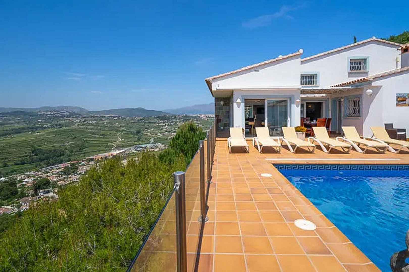 Terraza elevada con piscina de gresite azul, tumbonas blancas y barandilla de cristal con vistas panorámicas al valle y las montañas de Moraira.